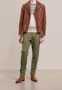 Hnědá nubuková motorkářská bunda se zipovými detaily, krémový svetr s barevným geometrickým vzorem, olivově zelené kalhoty a hnědé nubukové boty.