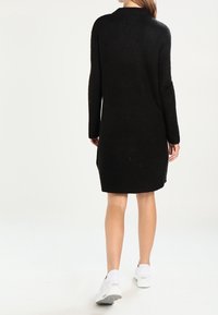Donna che indossa un vestito a maglia nero con maniche lunghe e sneakers bianche, in piedi contro uno sfondo bianco semplice, con la schiena rivolta verso la telecamera.