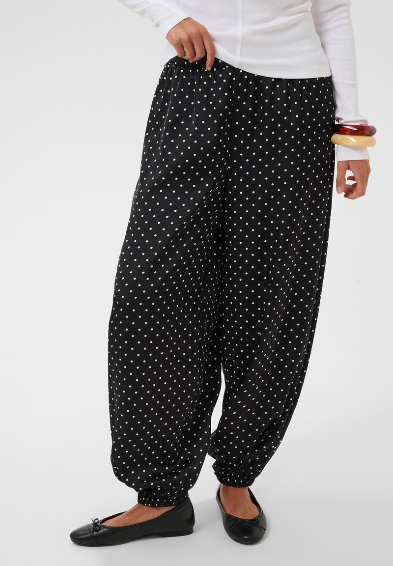 Personne portant un pantalon noir ample à pois blancs, taille et chevilles élastiques, associé à des ballerines noires et un haut blanc à manches longues.