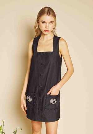 Femeie îmbrăcată într-o rochie neagră fără mâneci, cu nasturi și broderie florală pe buzunare, stând în fața unui fundal bej.