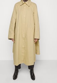 Beige oversized bomullskappa med spetsig krage, framknappar och sidofickor, matchad med svarta rutiga byxor och ankelskors.