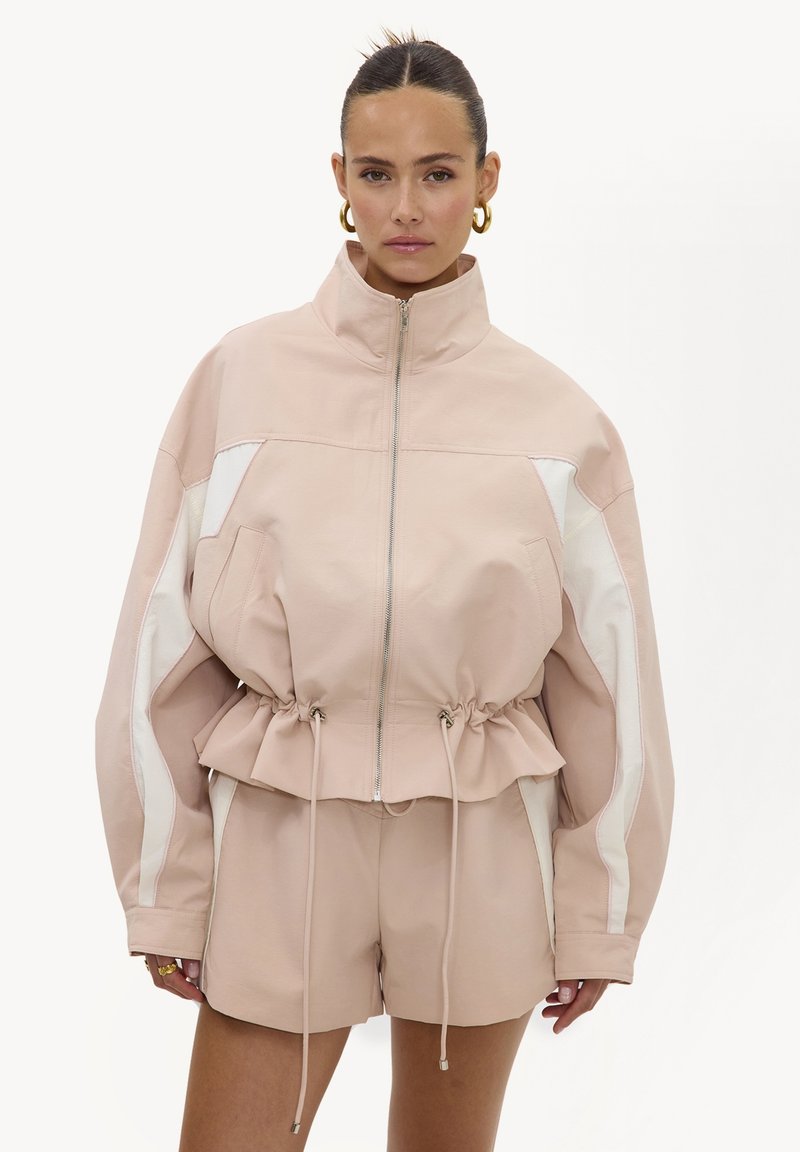 Femme portant une veste beige zippée avec des rayures blanches, taille cintrée, short assorti et boucles d'oreilles anneaux dorés, debout sur fond blanc.