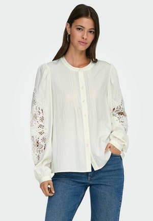 Camicia bianca a maniche lunghe con bottoni, con dettagli in pizzo sulle maniche, caratterizzata da pieghe sul davanti e un colletto arrotondato.