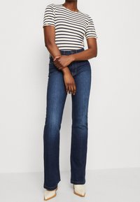 Randig kortärmad topp i kräm och marinblå, parat med mörkblå utsvängdajeans. Toppenn har en rund halsringning och figurnära design.