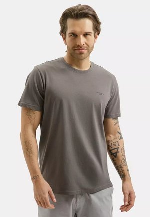 Mężczyzna z krótkimi brązowymi włosami, ubrany w szarą koszulkę i jasnoszare spodnie, pokazujący tatuaże na obu ramionach na jednolitym tle.