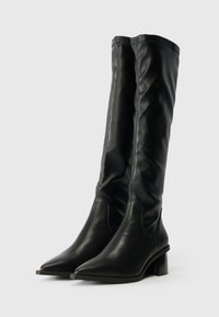 Fekete bőrből készült, térd felett érő csizmák, hegyes orral, diszkrét varrással és vastag sarkával. Simaság jellemzi, az elegáns, testhez simuló kialakítás mellett.