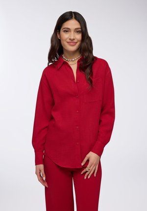 Camicia rossa con bottoni realizzata in un tessuto testurizzato, con colletto, maniche lunghe e taschino sul petto. Abbinata a pantaloni rossi coordinati.