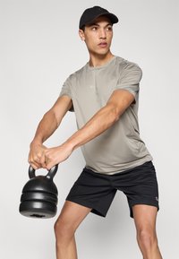 Αθλητική γκρι ανοιχτή μπλούζα και μαύρο σορτς. Το μοντέλο κρατάει μια μαύρη καμπάνα γυμναστικής, δείχνοντας μια λεπτή εφαρμογή και ύφασμα που απομακρύνει την υγρασία.