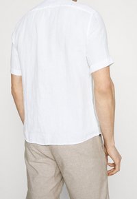 Camicia bianca in lino a maniche corte con colletto button-down, con polsini arrotolati e una vestibilità comoda, abbinata a pantaloni in lino beige chiaro.