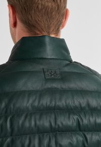 Dunkelgrüne Lederweste mit hohem Kragen, gestepptem Muster und einem kleinen Logo-Patch auf dem Rücken. Glatte Textur und taillierte Passform.