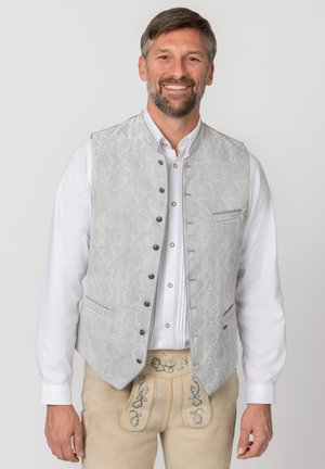 Lächelnder Mann mit weißem Hemd, hellgrauer gemusterter Weste mit schwarzen Knöpfen und beigen bestickten traditionellen Lederhosen.