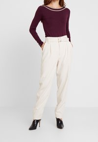 Burgundy pitkähihaisella puserolla, jossa on leveä kaula-aukko ja beige reunus, yhdistettynä korkeavyötäröisiin luonnonvalkoisiin housuihin ja mustiin korollisiin mokkasiineihin.