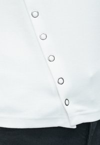 Biely rebrovaný top s striebornými kovovými očkami pozdĺž lemovania. Hladká textúra, so jemným asymetrickým dizajnom a priliehavým strihom.