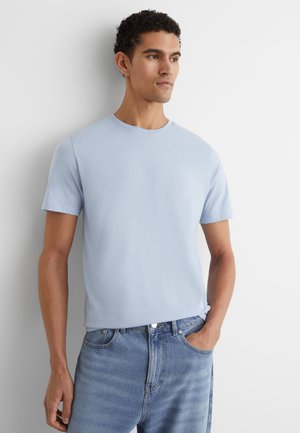 Mann mit kurzen lockigen Haaren, der ein hellblaues Kurzarm-T-Shirt und blaue Jeans trägt, steht mit einer Hand in der Tasche und schaut zur Seite.