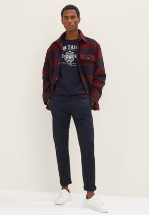 Rote und marineblaue Karoflanelljacke über einem marineblauen T-Shirt mit Grafik, kombiniert mit hochgekrempelten marineblauen Hosen und weißen Turnschuhen. Glatte Texturen und lässige Passform.