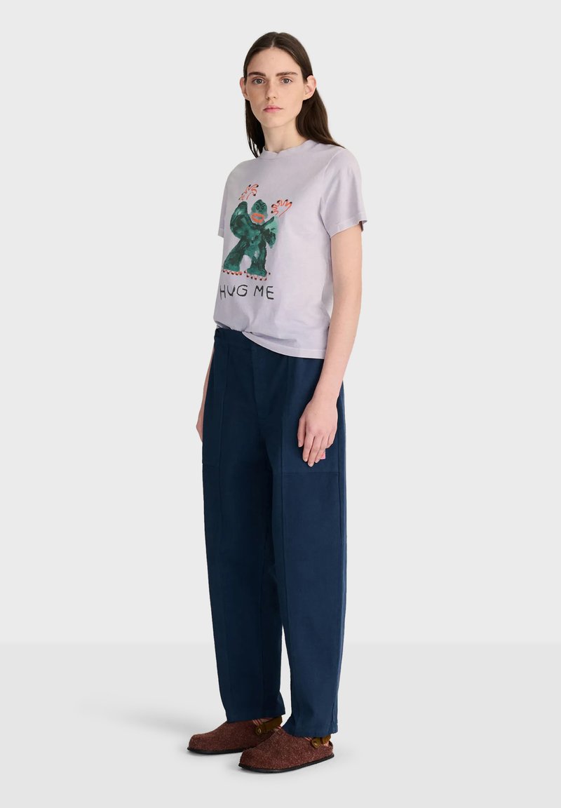 Jeune femme aux longs cheveux foncés portant un t-shirt violet clair avec un gorille graphique et le texte "HUG ME", un pantalon large bleu marine et des chaussures marron.