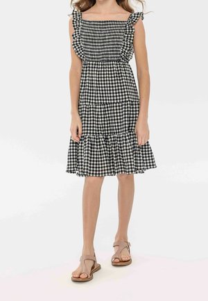 Pige iført en sort og hvid ternet ærmeløs kjole med flæser og beige åbne sandaler, stående på en hvid baggrund.