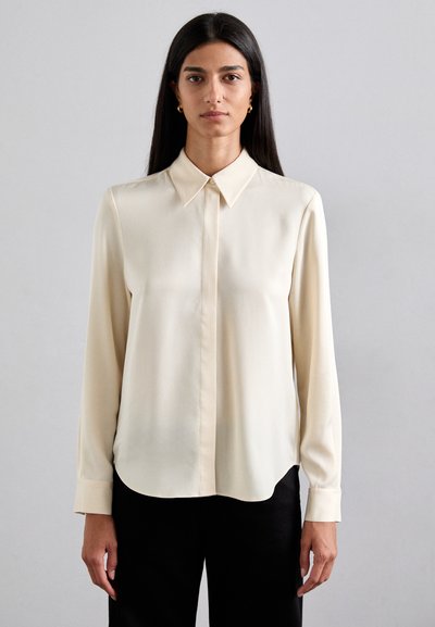Blouse crème en tissu léger et lisse avec un col classique, des boutons sur le devant et des manches longues. Bas arrondi.