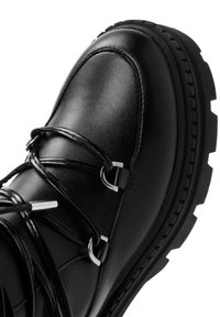 Botín de cuero negro con cordones, con herrajes plateados, suela de goma y superficie texturizada. Presenta una puntera redonda y un diseño robusto.