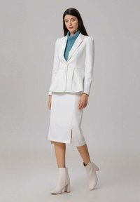 Femme debout portant un blazer blanc et une jupe midi assortie, un pull col roulé teal, et des bottines rose clair sur un fond gris.