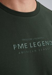 Tmavě zelené bavlněné tričko s kulatým výstřihem. Obsahuje velký plastický nápis "PME LEGEND" a "FREEDOM UNLIMITED" v světlejší zelené barvě.