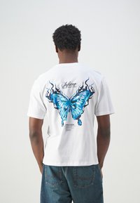 Witte katoenen t-shirt met een grote blauwe vlinderafbeelding met zwarte vlamaccenten op de achterkant. Korte mouwen en een relaxte pasvorm.
