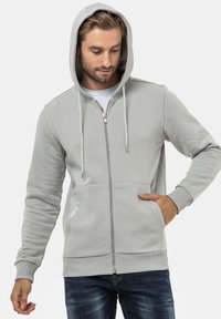 Sweat à capuche zippé gris avec cordon de serrage, poignets côtelés et poches avant. Tissu lisse avec une texture douce.