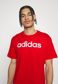 Sarkans kokvilnas T-krekls ar izteiksmīgu baltu "adidas" logo uz krūšu daļas. Īsas piedurknes, apaļa kakla izgriezums, gluda tekstūra.
