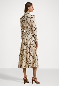 Long-sleeved dress in cream fabric featuring a brown chain pattern, cinched waist, and a flowing skirt. Accented with heeled sandals.