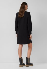 Schwarzes Kleid mit langen Puffärmel und seitlichen Schlitzen, aus einem glatten Stoff gefertigt. Getragen mit Stiefeletten mit einer massiven Sohle und elastischen Seitenpaneelen.