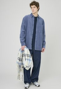 Chemise en velours côtelé bleu à boutons portée sur un haut bleu marine, associée à un pantalon en denim bleu ample. Tenant une écharpe douce en plaid de couleurs claires.