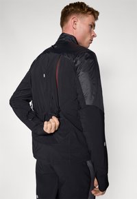 Schwarze Sportjacke mit einer Mischung aus matten und glänzenden Texturen, sichtbaren Reißverschlüssen, hohem Kragen und verstellbaren Elementen auf der Rückseite.