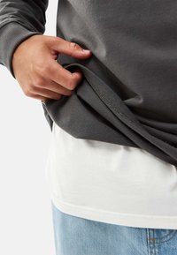 Gray sweatshirt layered over a white t-shirt, showing soft fabric texture and visible stitching with a hand grasping the hem. Blue jeans visible below.