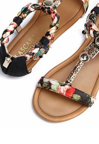 Zwei Sandalen mit einem geflochtenen Blumenriemen in Schwarz, geschmückt mit bunten Akzenten und metallischen Beschlägen. Glattes, sandfarbenes Fußbett und strukturierte Laufsohle.