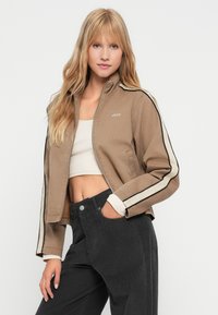 Jovem mulher com cabelo loiro comprido, usando uma jaqueta bege com zíper e riscas laterais, uma blusa branca cropped e calças pretas de cintura alta, com a mão no bolso.