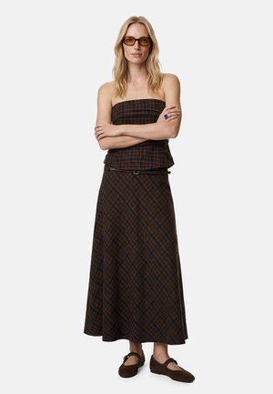 Mujer con cabello rubio que lleva gafas de sol broncíneas, top de cuadros marrón sin tirantes y falda larga a juego con zapatos marrones, brazos cruzados.