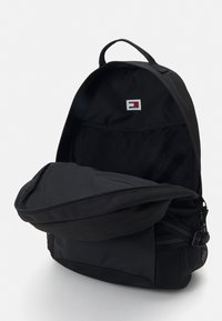 Mochila negra con un compartimento principal con cremallera, bolsillos de malla a los lados y un pequeño parche con el logo en el panel posterior interior. Textura suave.
