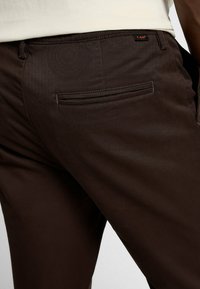 Brązowe spodnie wykonane z gładkiego materiału, z tylną kieszenią z subtelnym przeszyciem i małą metką z logo. Klasyczny krój.