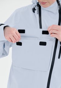 Ljusgrå vattentät jacka med justerbar huva, framfickor med kardborre och svarta detaljer. Har en elegant och modern design.