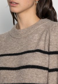 Beige Wollpullover mit schwarzen horizontalen Streifen, ausgestattet mit einem runden Halsausschnitt und gerippten Bündchen, der eine weiche und strukturierte Oberfläche zeigt.