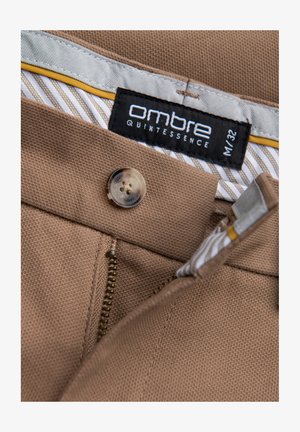 Braune Stoffhose mit Reißverschluss und Knopfverschluss. Das Etikett trägt die Aufschrift "Ombre Quintessence, M/32." Das Futter hat ein gestreiftes Muster in hellen Farben.