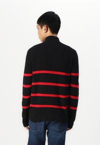 Črn rebrast pulover s horizontalnimi rdečimi črtami, visokim ovratnikom in dolgimi rokavi, združen z modrimi jeans hlačami.
