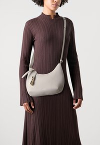 Borsa a tracolla in pelle grigia con forma curva, hardware dorato e chiusura con zip, indossata sopra un vestito marrone a coste con scollatura alta.