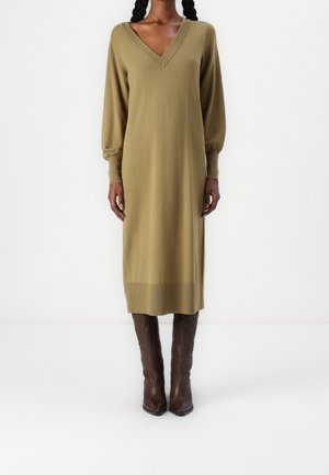 Woman wearing olive green long-sleeve V-neck midi knit dress paired with knee-high brown leather boots, standing against a white background.