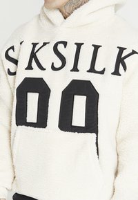 Fleece-Kapuzenpullover in Off-White mit schwarzem "SKSILK" Schriftzug und großer "00" Zahl auf der Vorderseite. Verfügt über eine Kängurutasche und eine strukturierte Oberfläche.