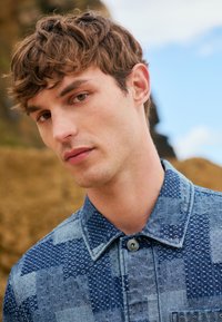 Giovane uomo con capelli corti e ricci castani che indossa una camicia di jeans blu con motivo, in piedi all'aperto con sfondo naturale sfocato.