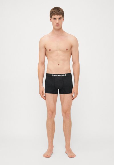 Black boxer briefs feature a repeating logo pattern, elastic waistband with "DSQUARED2," and a snug fit. Model poses barefoot, no shirt.