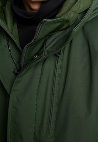 Chaqueta acolchada verde con cierre de cremallera, capucha ajustable y dos bolsillos frontales. Tejido texturizado con un acabado suave y costuras visibles.