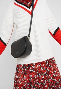 Borsa a tracolla nera con hardware dorato indossata sopra un maglione di maglia bianco con accenti rossi e neri e una gonna floreale rossa.
