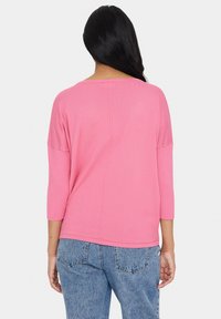 Rozā, viegla, adīta blūze ar trīs ceturtdaļu piedurknēm, brīvu piegriezumu, teksturētu apdari un centrālo šuvju detaļu aizmugurē. Valkāta ar zilām džinsām.